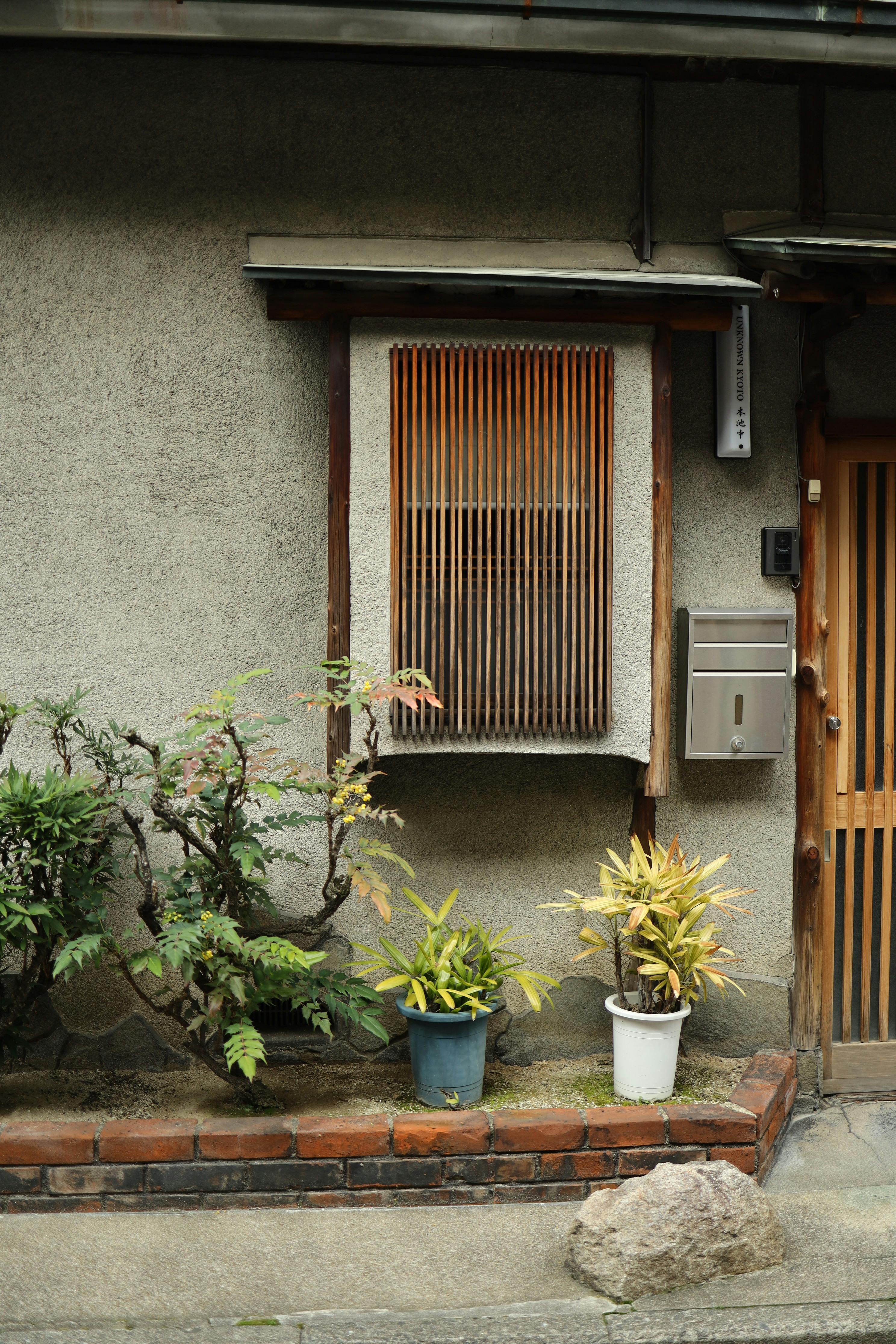A serene view of a traditional Japanese house facade adorned with potted plants and a wooden window shutter.