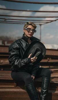 Stylish individual wearing black leather jacket, holding a hat, sitting outdoors with a modern vibe.