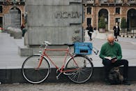 Red Bike Parked Near Pathway And A Man With His Dog Reading Book While Sitting