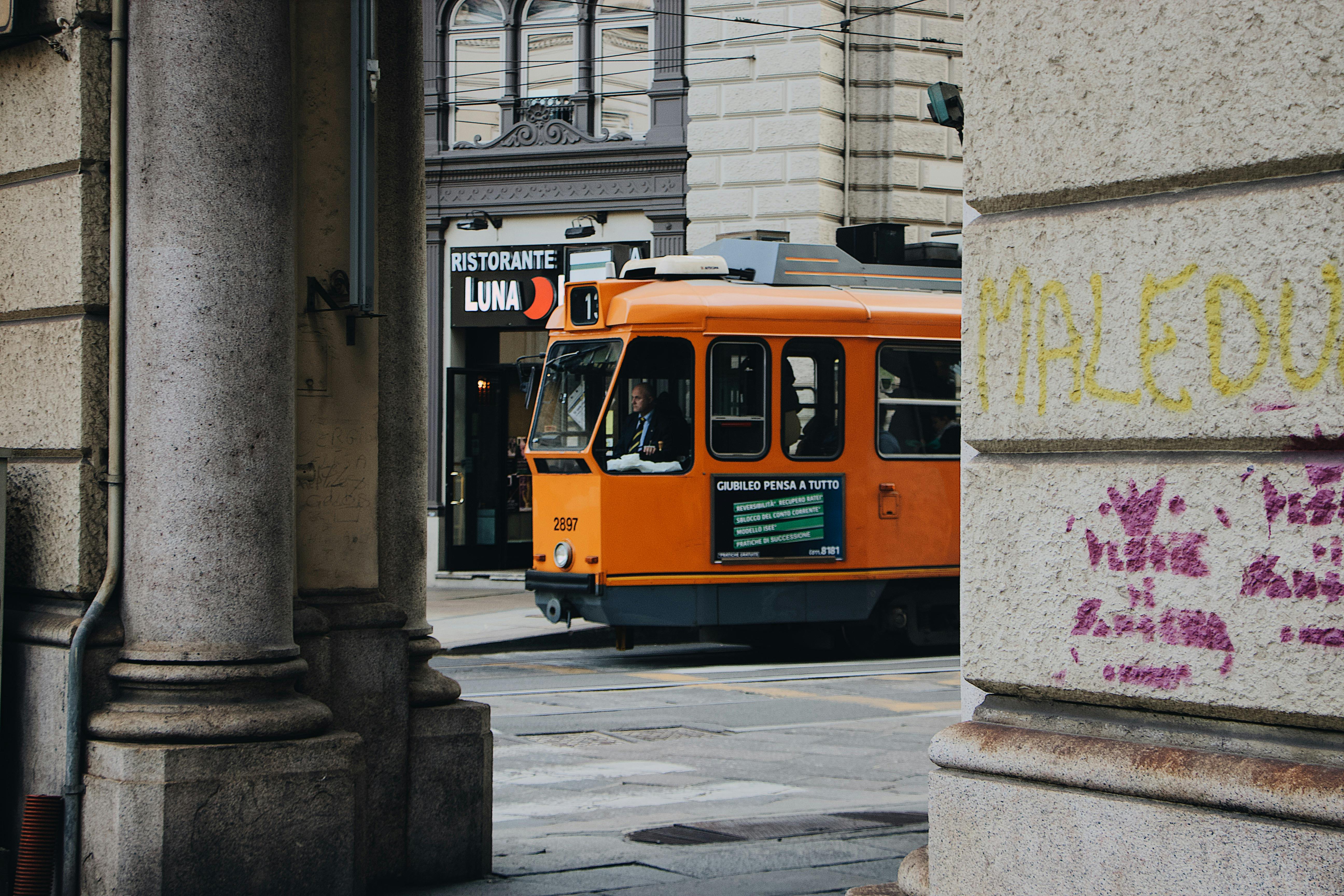 一辆老式橙色电车穿过历史悠久的城市街道，街道两旁建筑林立，还有意大利餐厅的招牌