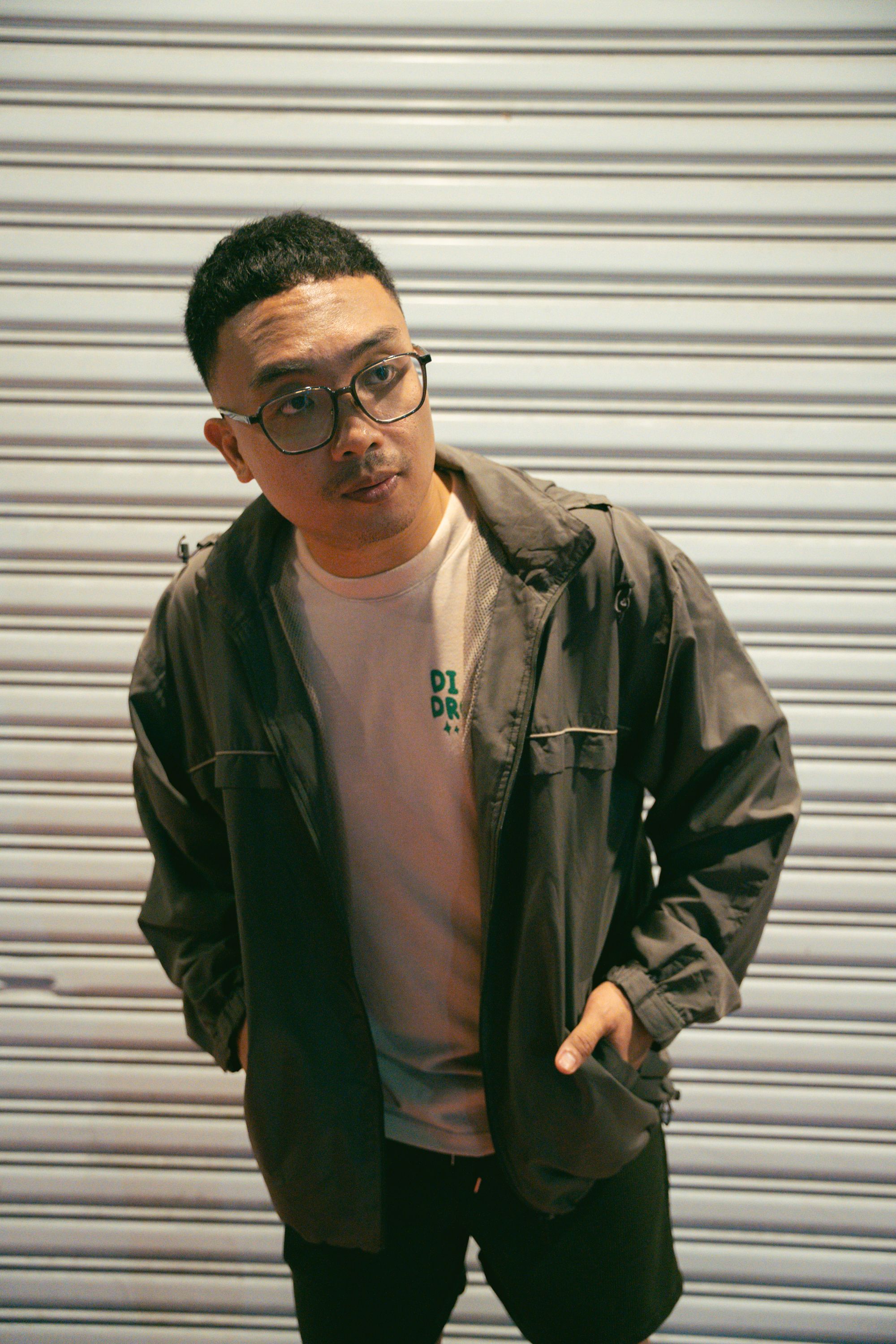 A confident young man in glasses stands against a city shutter, exuding a relaxed urban style.