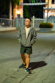 A young man strolling along a city street at night, wearing a jacket and shorts.