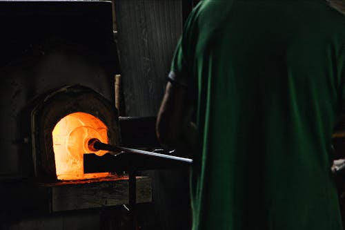 Free Back view of crop unrecognizable professional worker standing in front of furnace while blowing glass in workshop Stock Photo