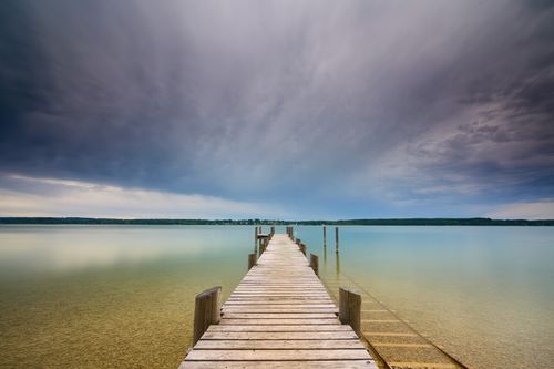 Free A tranquil view of a wooden pier extending into the serene Starnberger See under a dramatic sky. Stock Photo