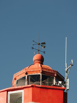 晴朗的蓝天下，红色灯塔设有风向标和天线，突显其航海导航功能