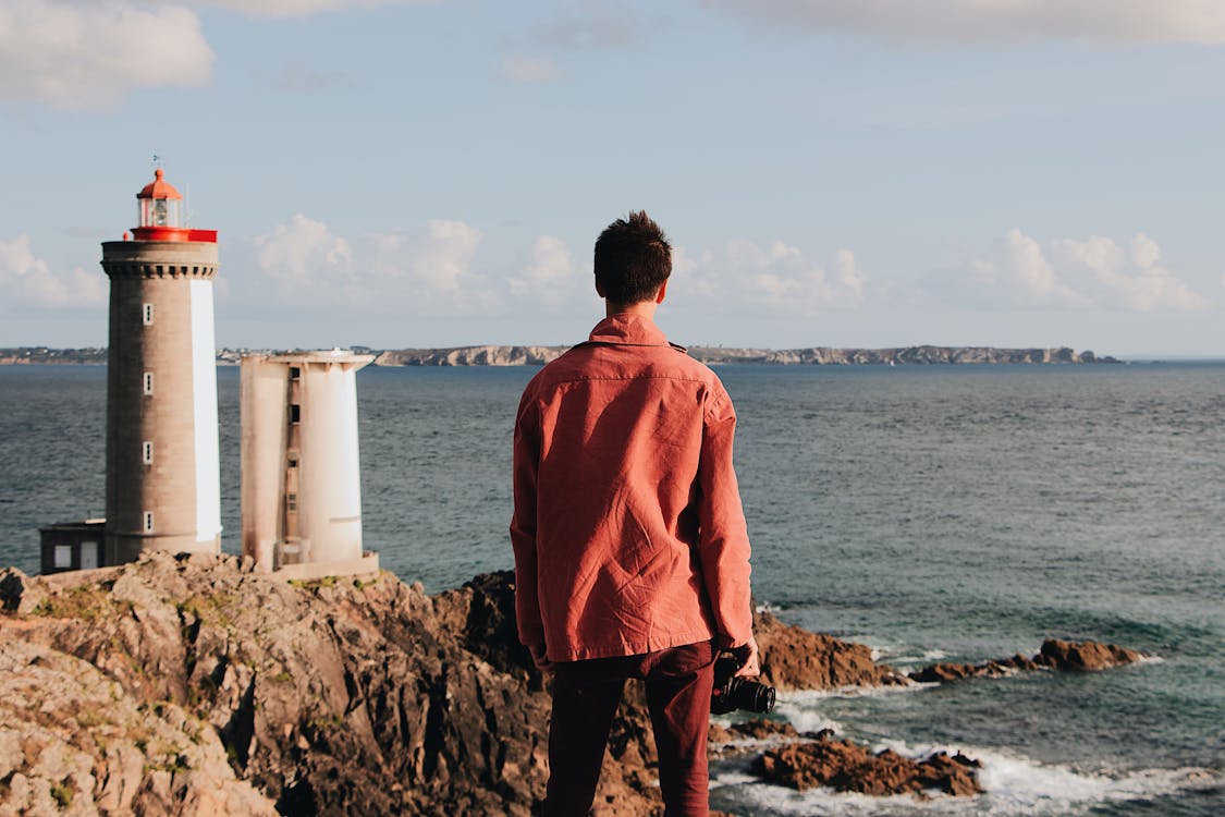 Free Back view of unrecognizable male traveler in casual red clothes contemplating seascape while standing on rocky shore with lighthouse near rippling sea Stock Photo