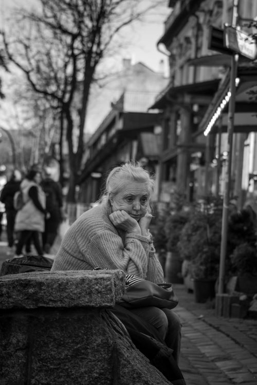 Free Black and white photo of an elderly woman sitting thoughtfully on a city street. Stock Photo