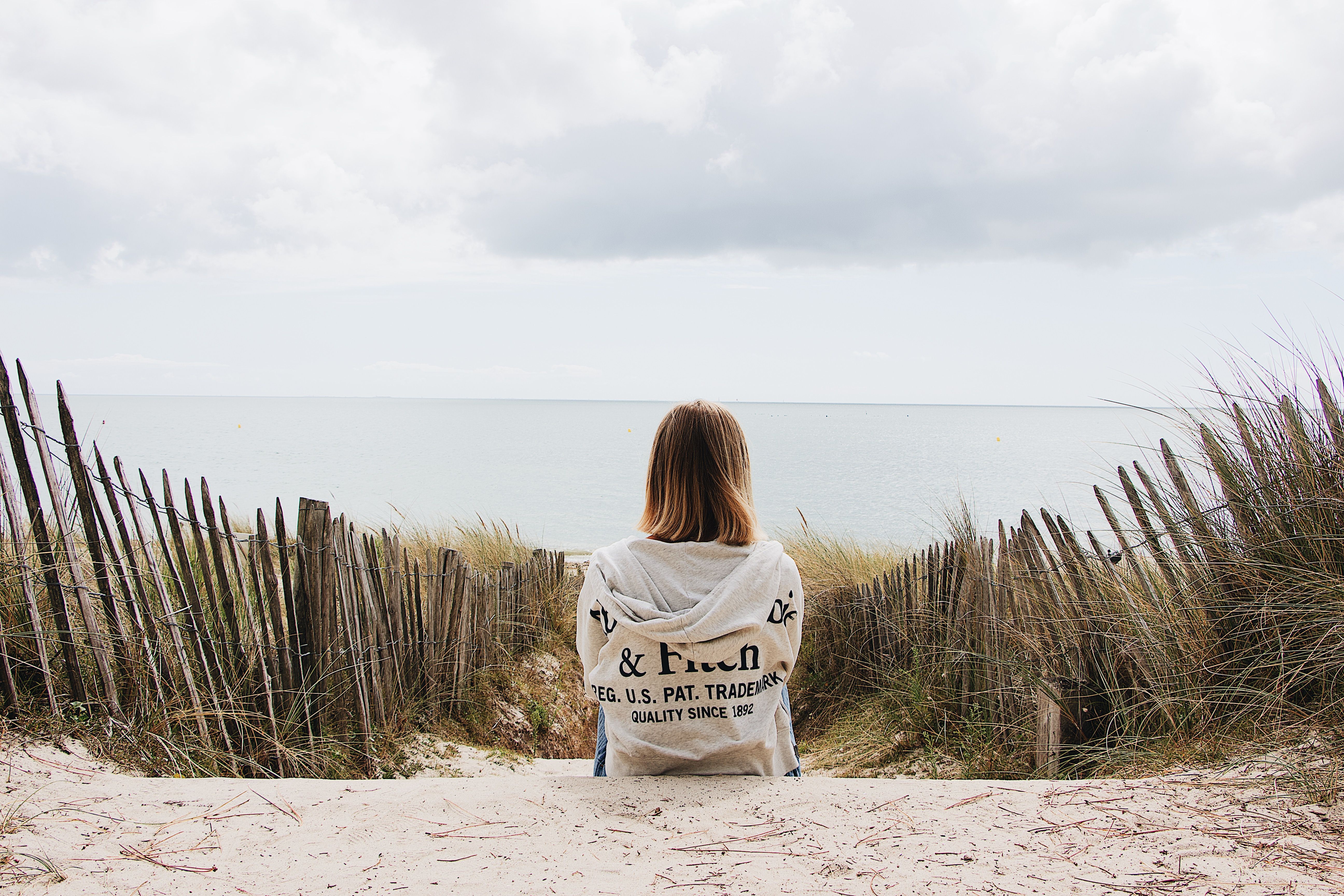 A woman sitting on a sandy path overlooking the ocean, surrounded by dunes and a rustic fence.