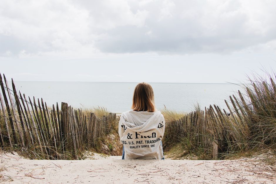 A woman sitting on a sandy path overlooking the ocean, surrounded by dunes and a rustic fence.