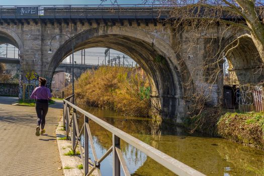 A lone jogger in sportswear runs along a serene canal under a historic stone bridge.