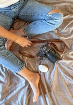 Casual indoor scene of a photographer relaxing on a bed with a camera and skincare product, wearing jeans.