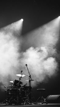 A dramatic black and white image of a drum set on stage with spotlights and smoke