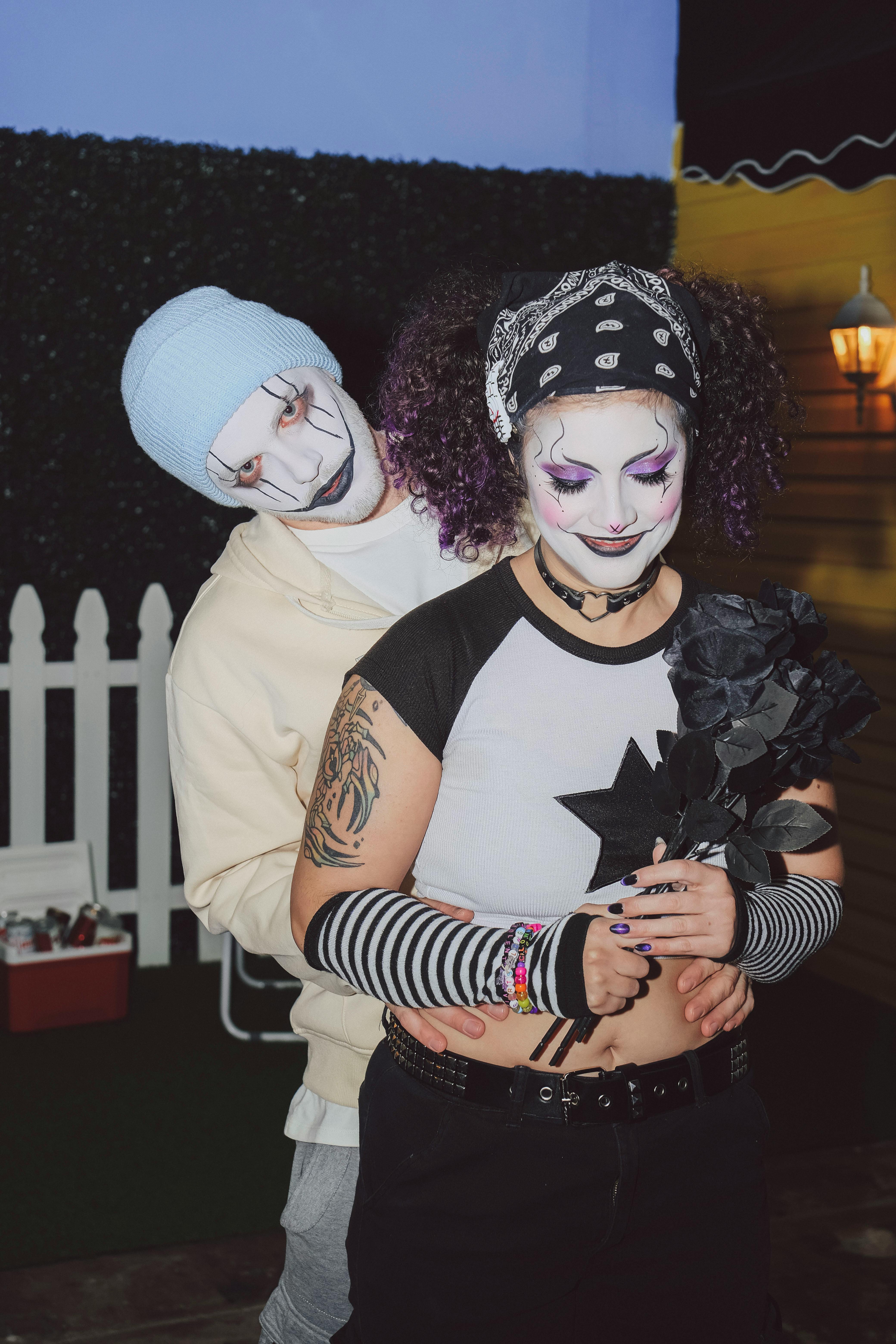 A gothic-styled couple in face paint and costumes share a tender embrace indoors.