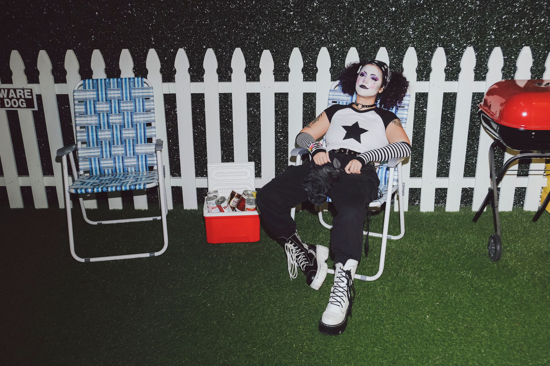 A person in gothic attire relaxes outdoors near a grill and cooler. Night setup.