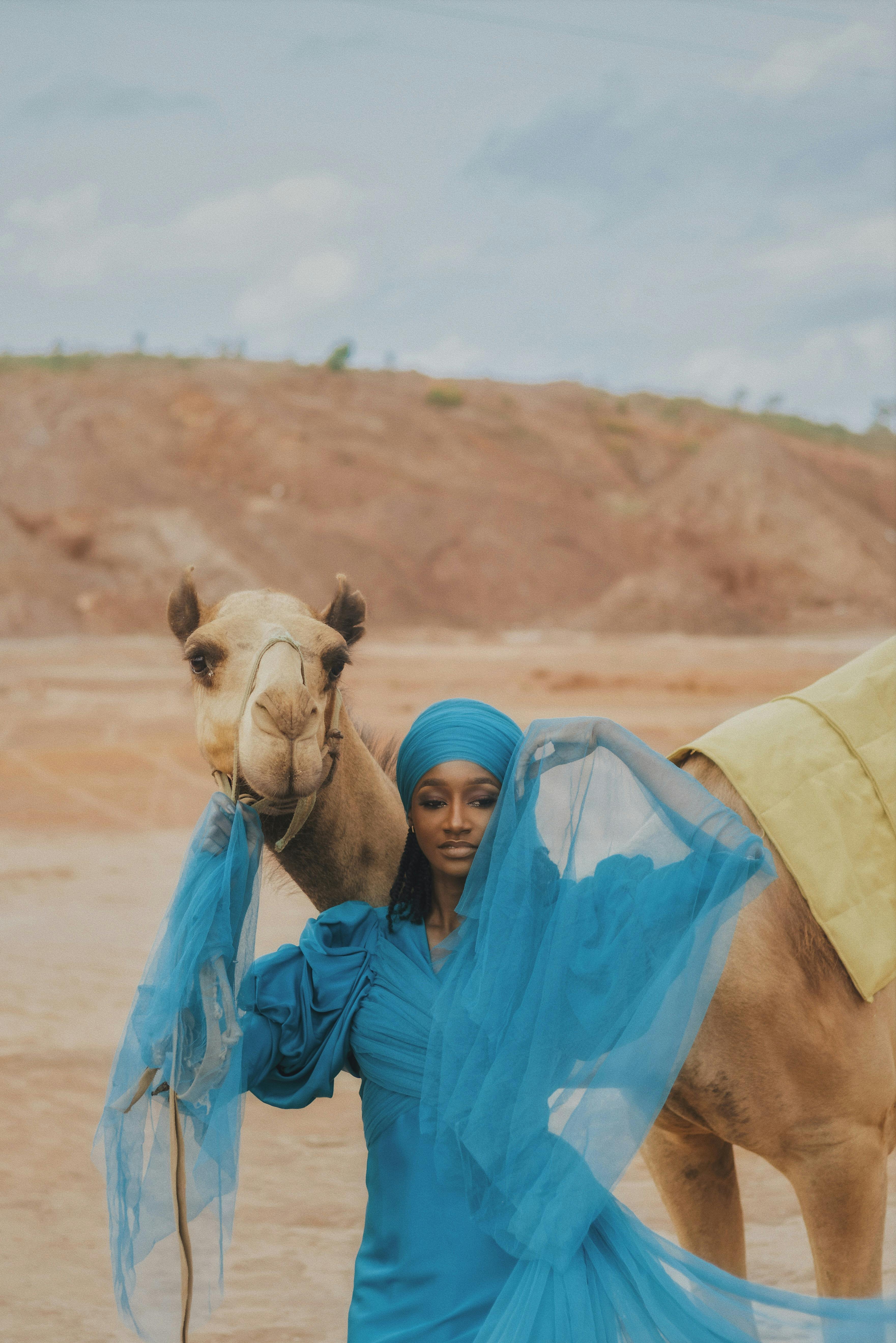 A woman in blue traditional attire poses with a camel in a desert landscape in Nigeria.