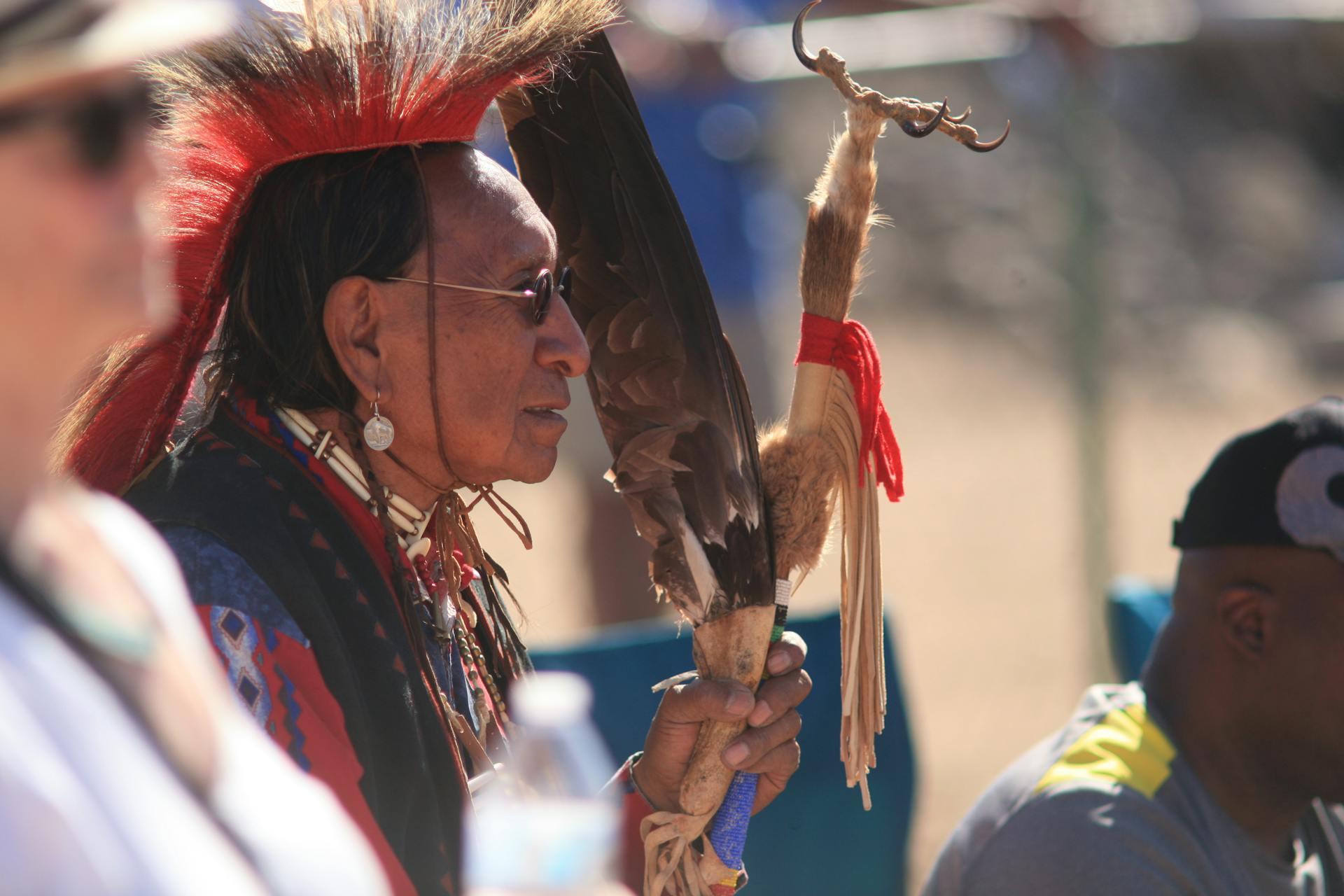 Native American elder in traditional attire holding a ceremonial staff outdoors. Captivating cultural representation.