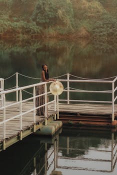 A woman in a sundress stands on a dock in Abuja, overlooking calm waters amid lush greenery.