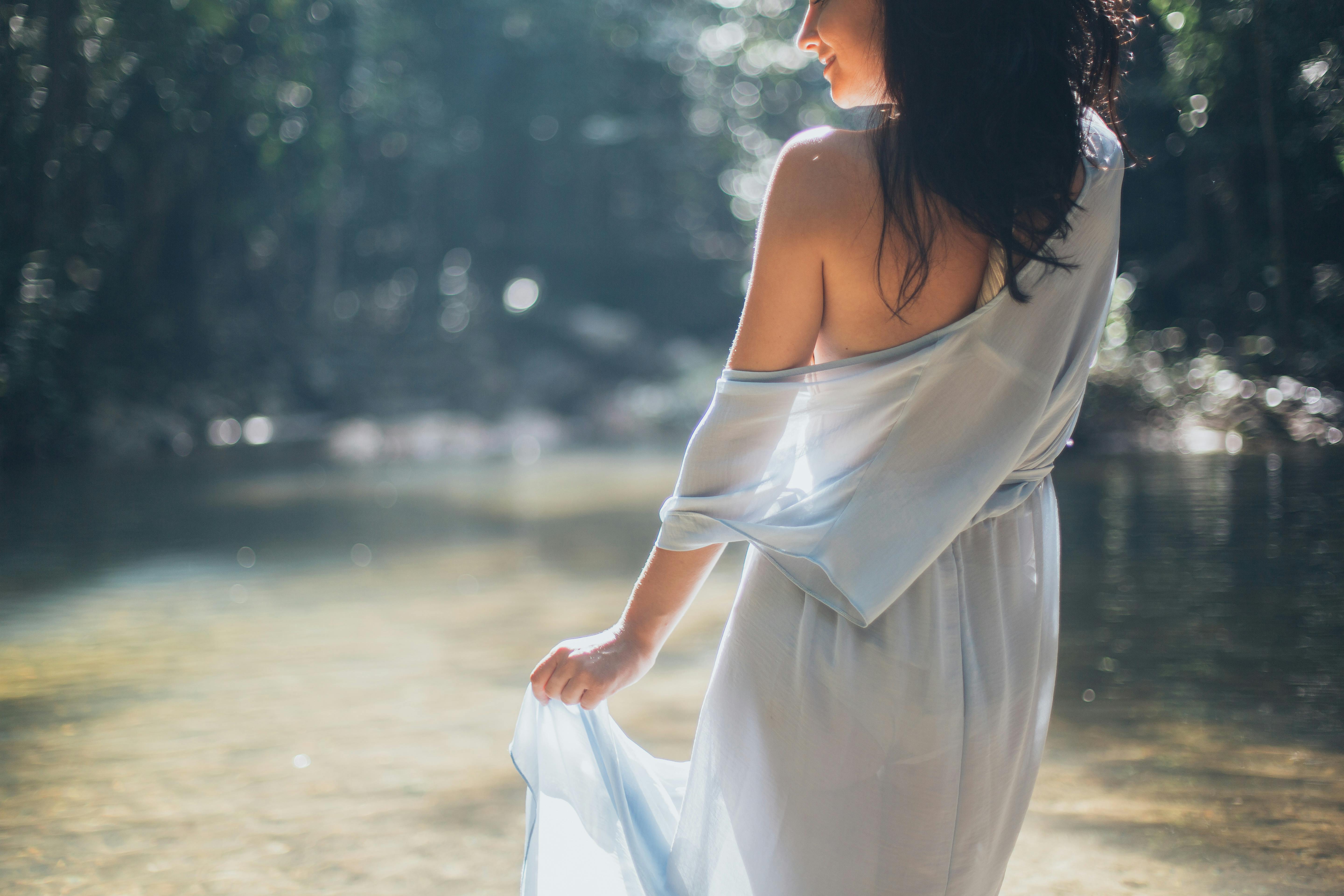 一位身着飘逸蓝色连衣裙的优雅女人，站在阳光明媚的森林里一条宁静的河边