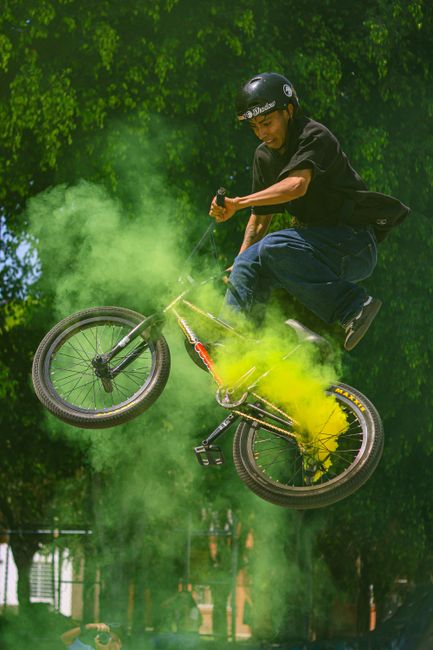 A BMX rider performs an aerial stunt with colorful smoke in an urban park.