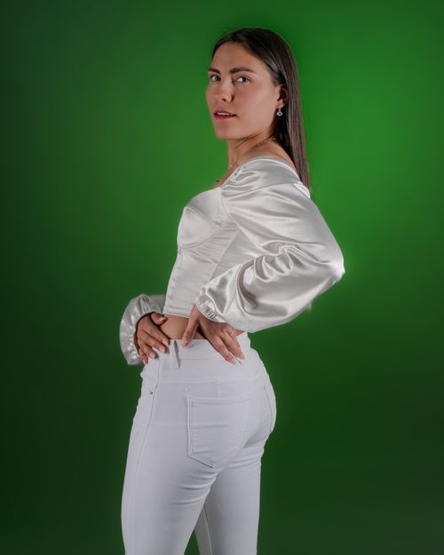 Free Portrait of a confident woman posing elegantly in a white satin top and pants against a vibrant green backdrop. Stock Photo