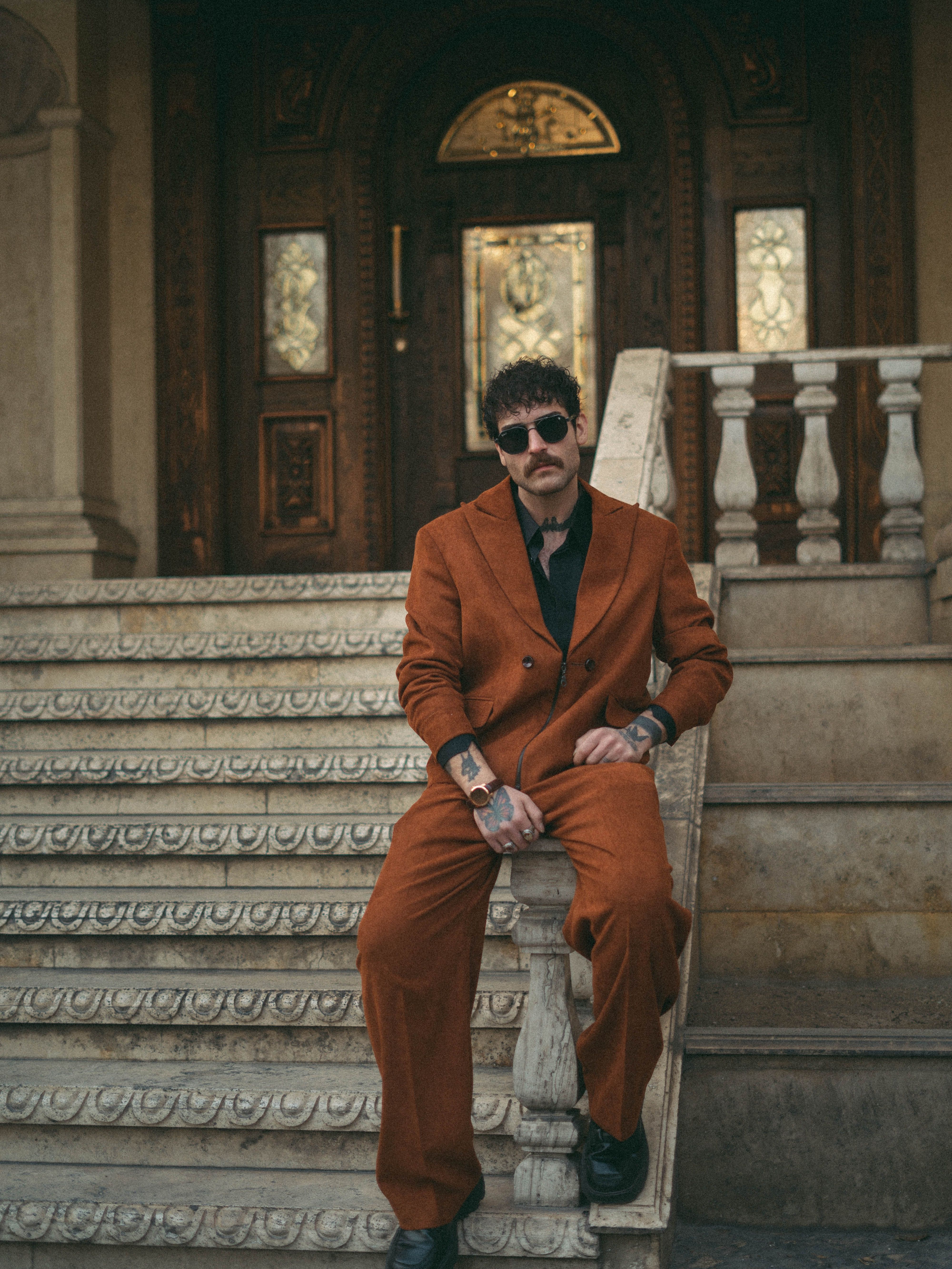 Fashionable man in an orange suit poses confidently on ornate outdoor staircase.