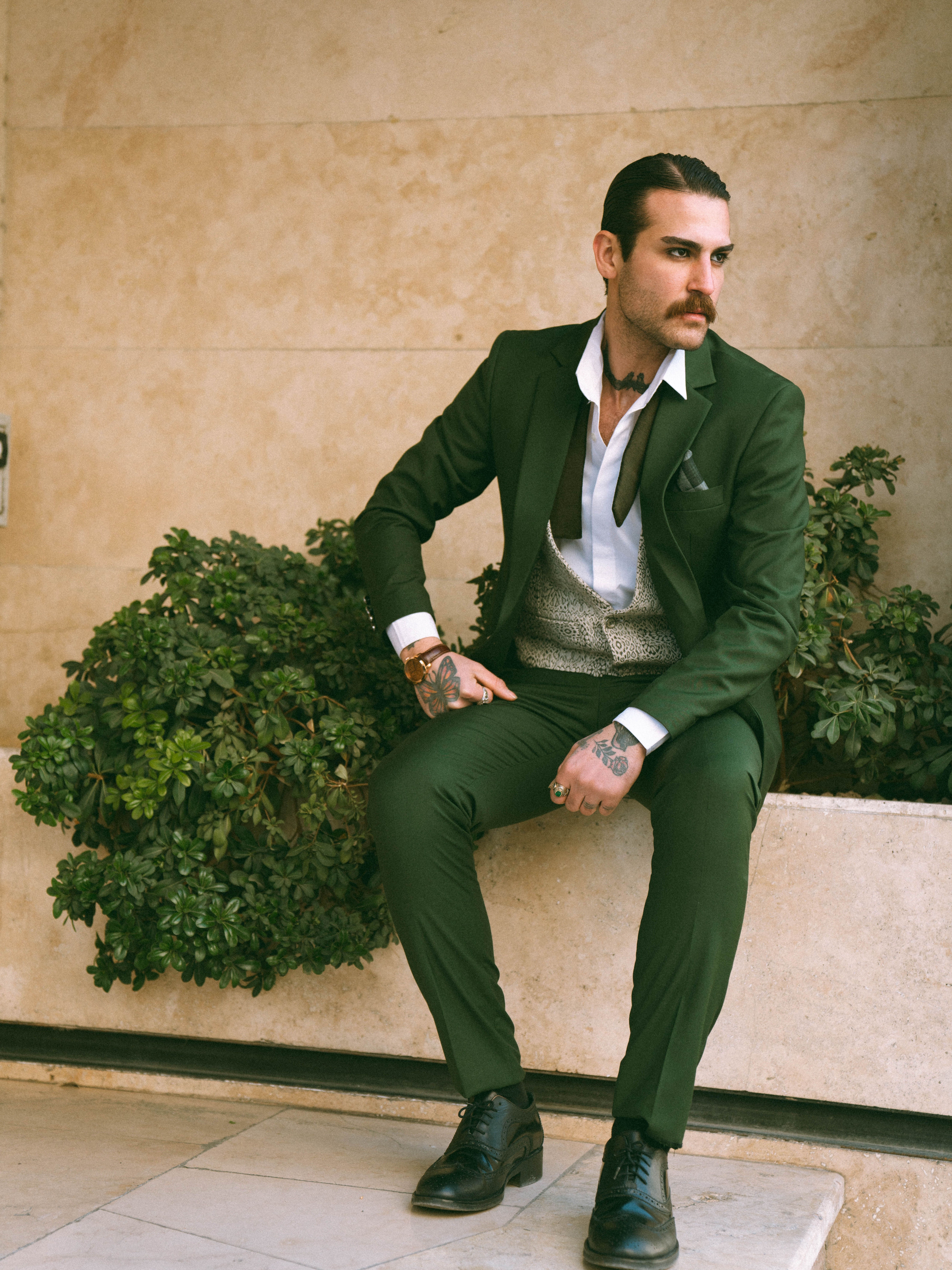A fashionable man in a green suit sitting outdoors next to plants, showcasing tattoos.