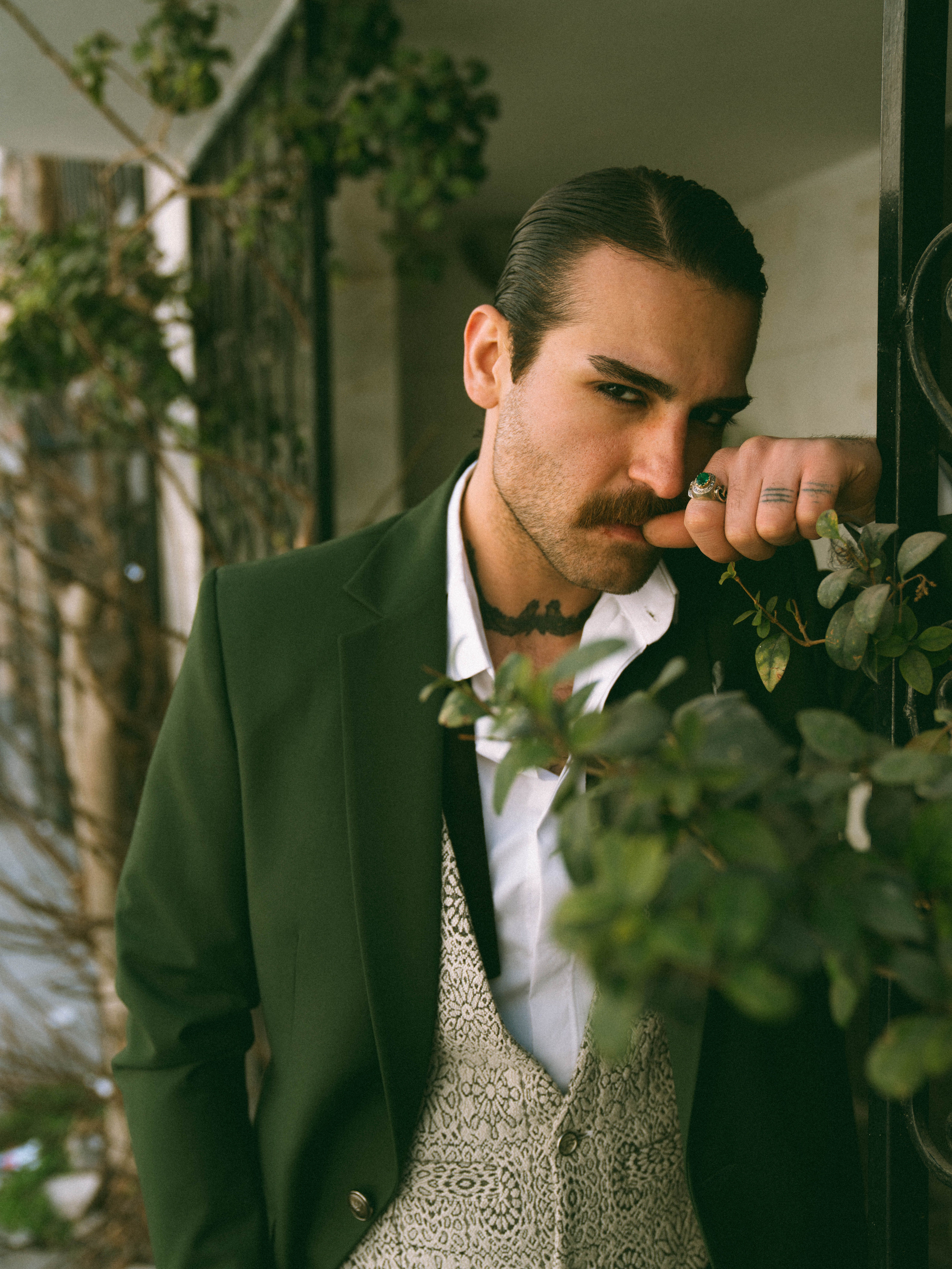 Man with mustache and tattoos poses outdoors in a stylish suit with greenery.