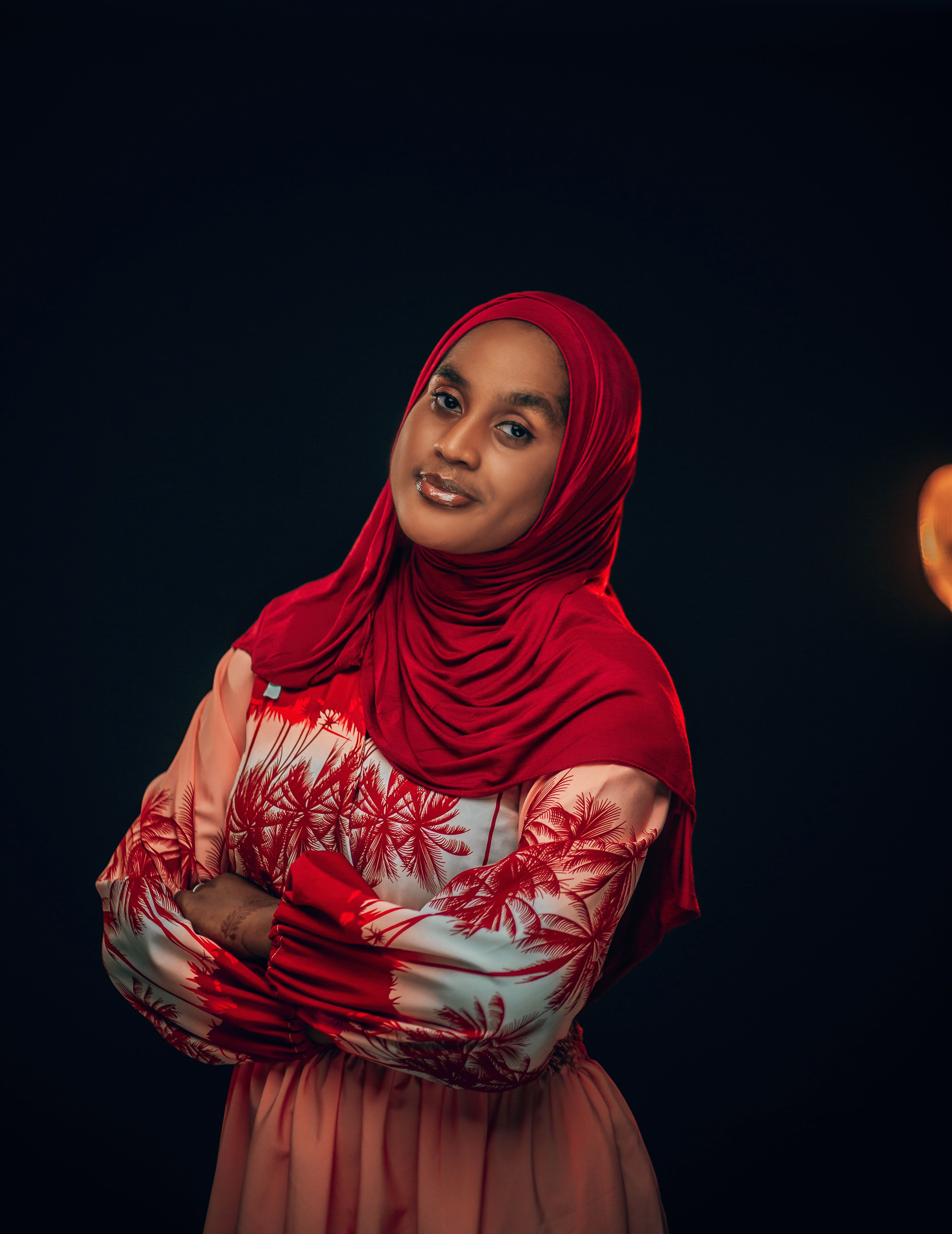 Studio portrait of a woman in a red hijab and floral dress, confidently posing with folded arms.