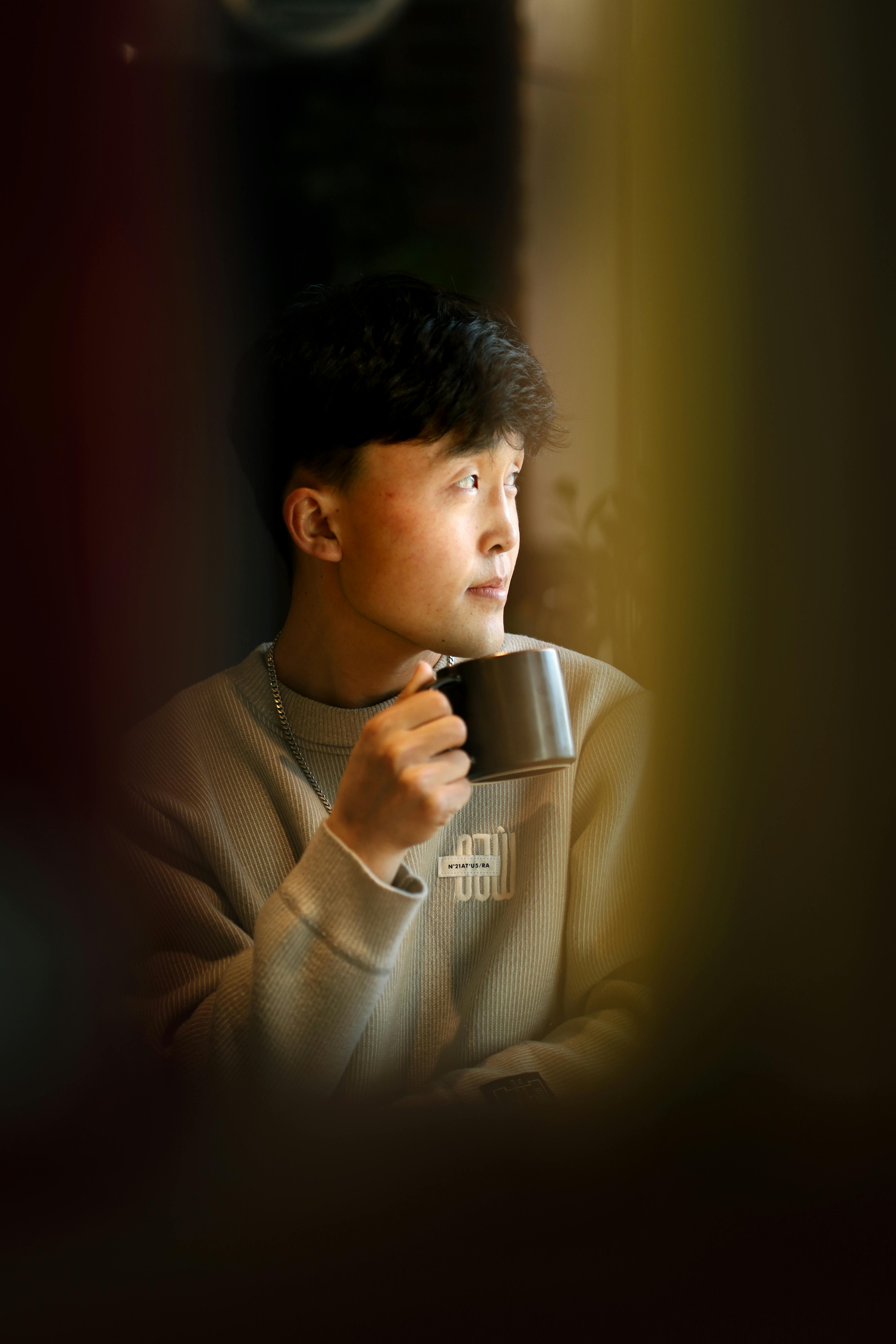 Gratis Un giovane si gode un momento di tranquillità sorseggiando un caffè in casa, immerso in una luce soffusa. Foto a disposizione