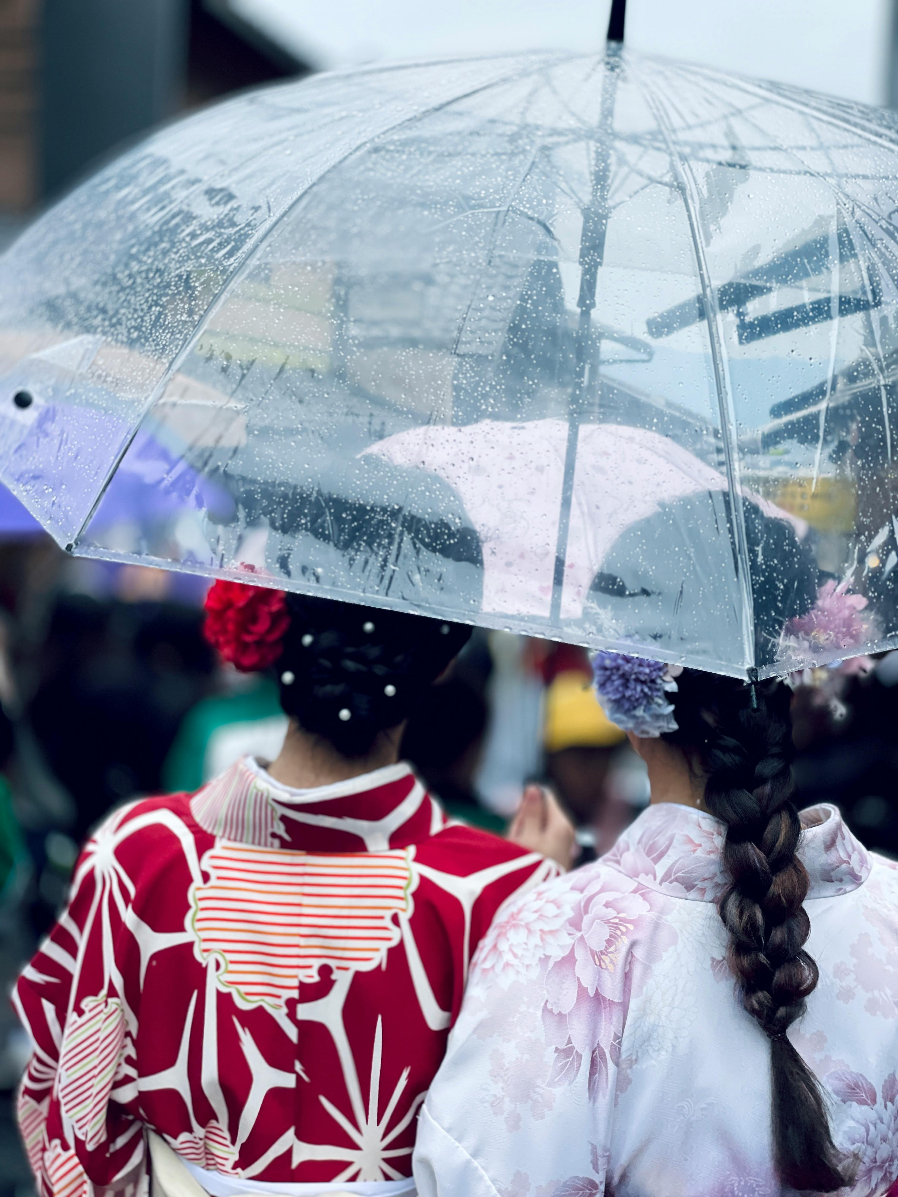 Two women in traditional kimonos walk under a clear umbrella during rain in Kyoto's vibrant street.