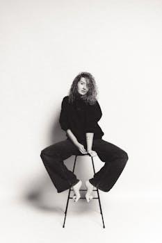 Artistic black and white portrait of a woman sitting on a stool, showcasing modern fashion.