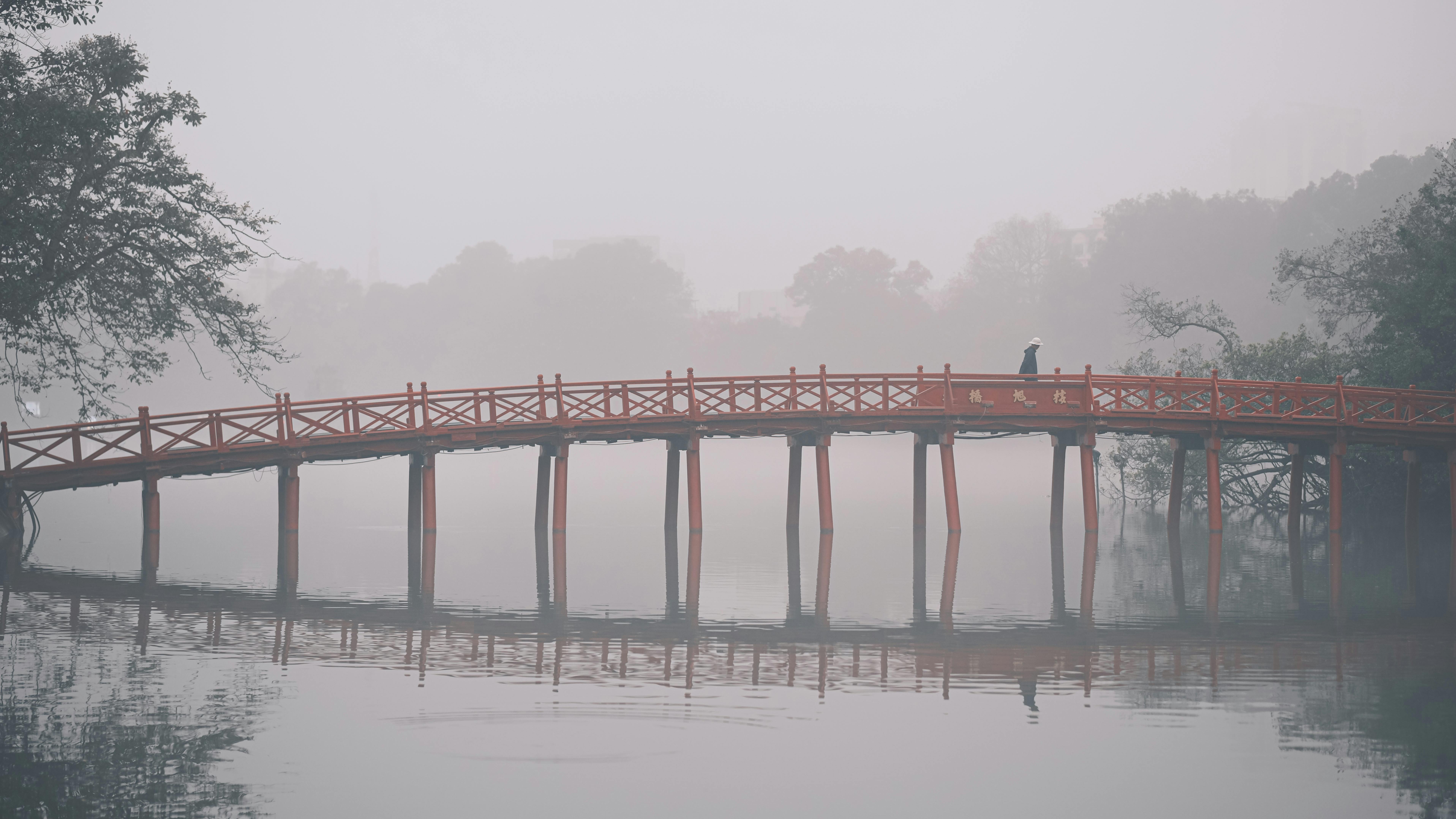 A person walking on a misty red bridge over calm waters with trees in the background.