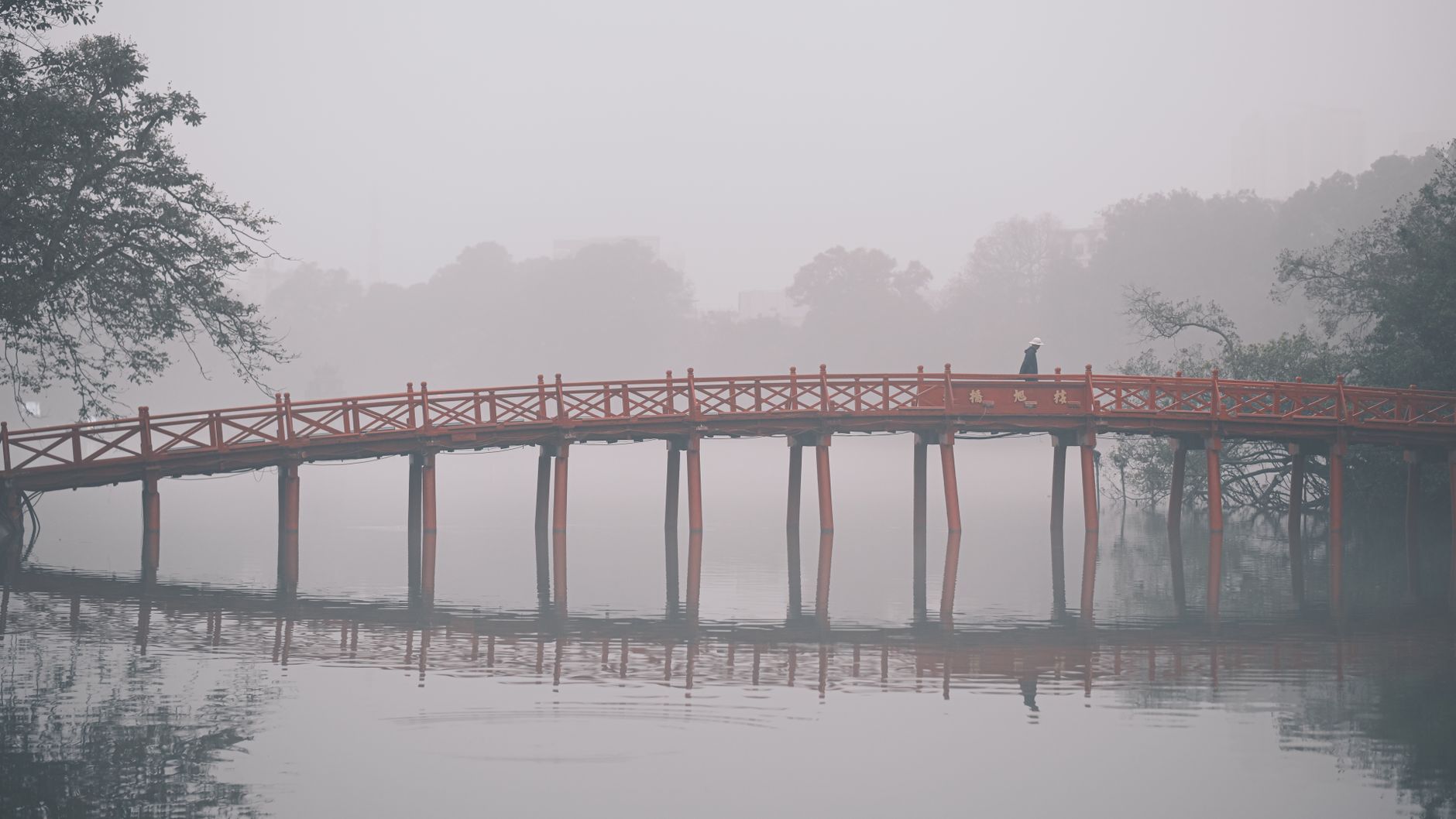 A person walking on a misty red bridge over calm waters with trees in the background.