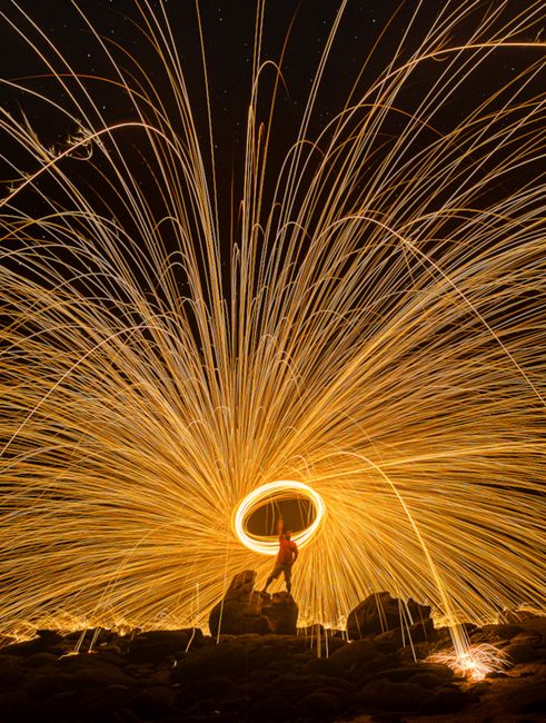 Fotomural Deslumbrante fotografía nocturna de larga exposición de fuego giratorio que crea estelas de luz sobre terreno rocoso.