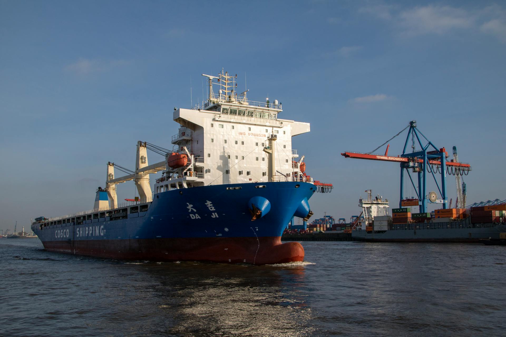 Cargo ship navigating the Elbe River in Hamburg harbor under a clear sky.