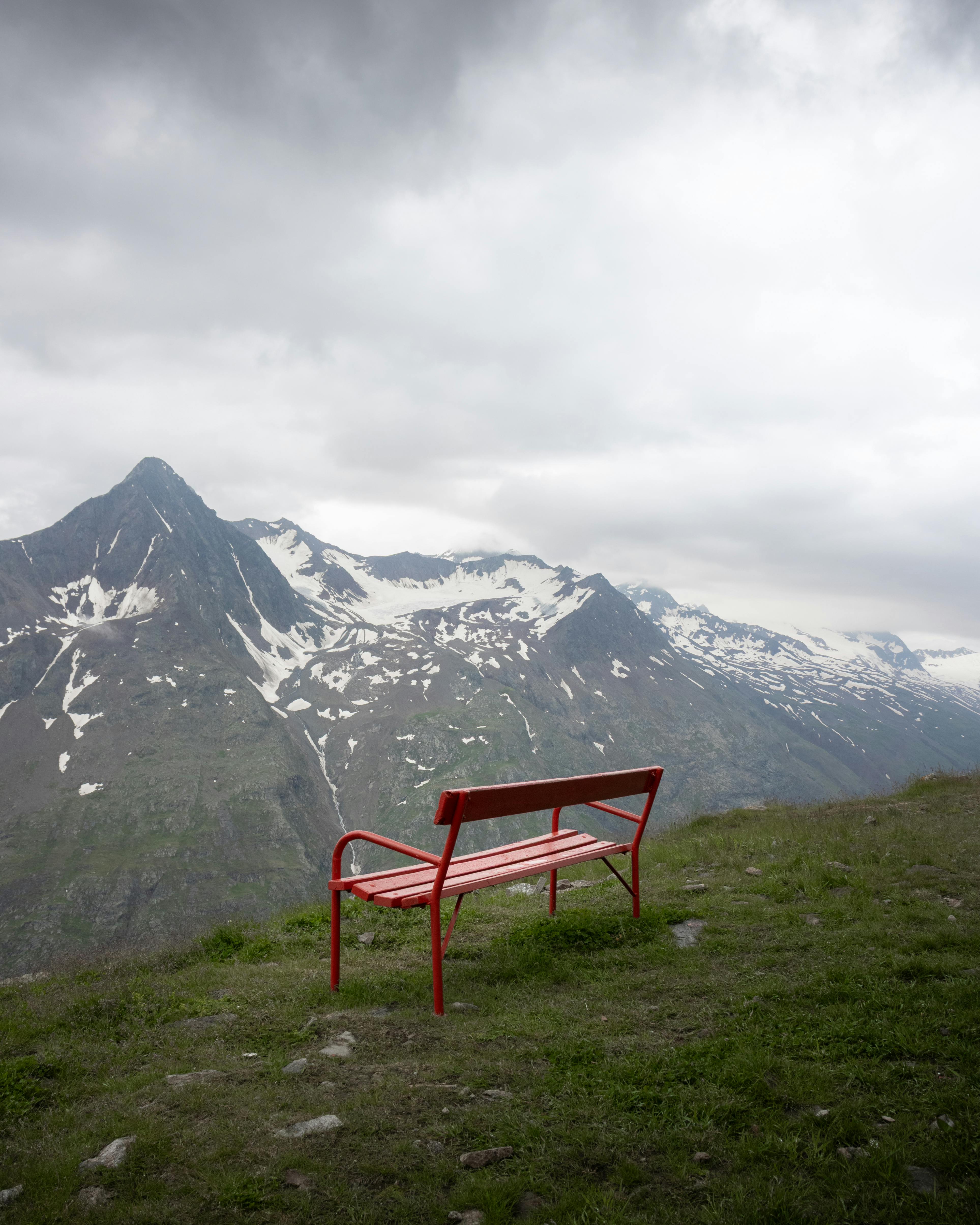 A lonely red bench on a grassy hilltop in Tirol offers stunning views of snow-capped Austrian Alps.