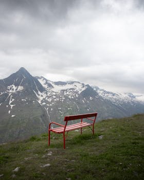 A lonely red bench on a grassy hilltop in Tirol offers stunning views of snow-capped Austrian Alps.