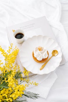 Delicate pastry paired with a golden spoon, mimosa flowers, and coffee on a white setting, exuding elegance.