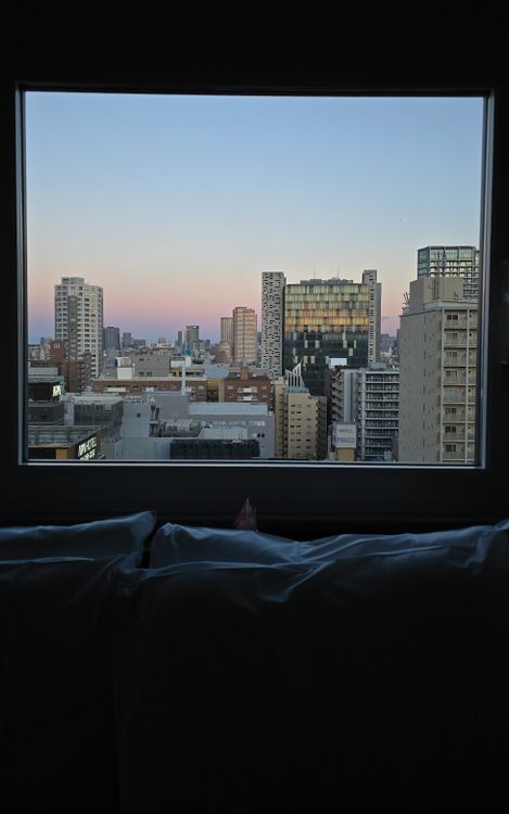 View from a dark bedroom bed toward a large window framing a mid-rise city skyline at dusk with a pink-blue sky. View from a dark bedroom bed toward a large window framing a mid-rise city skyline at dusk with a pink-blue sky.