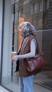 Stylish woman in hijab holding coffee and bag on an urban street, side view.