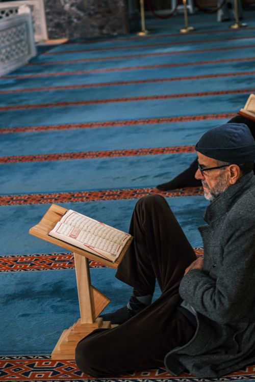 Free An elderly man reads the Quran in a tranquil mosque, surrounded by ornate carpets. Stock Photo