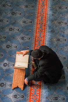 A man in traditional attire sits on a mosque carpet reading the Quran.