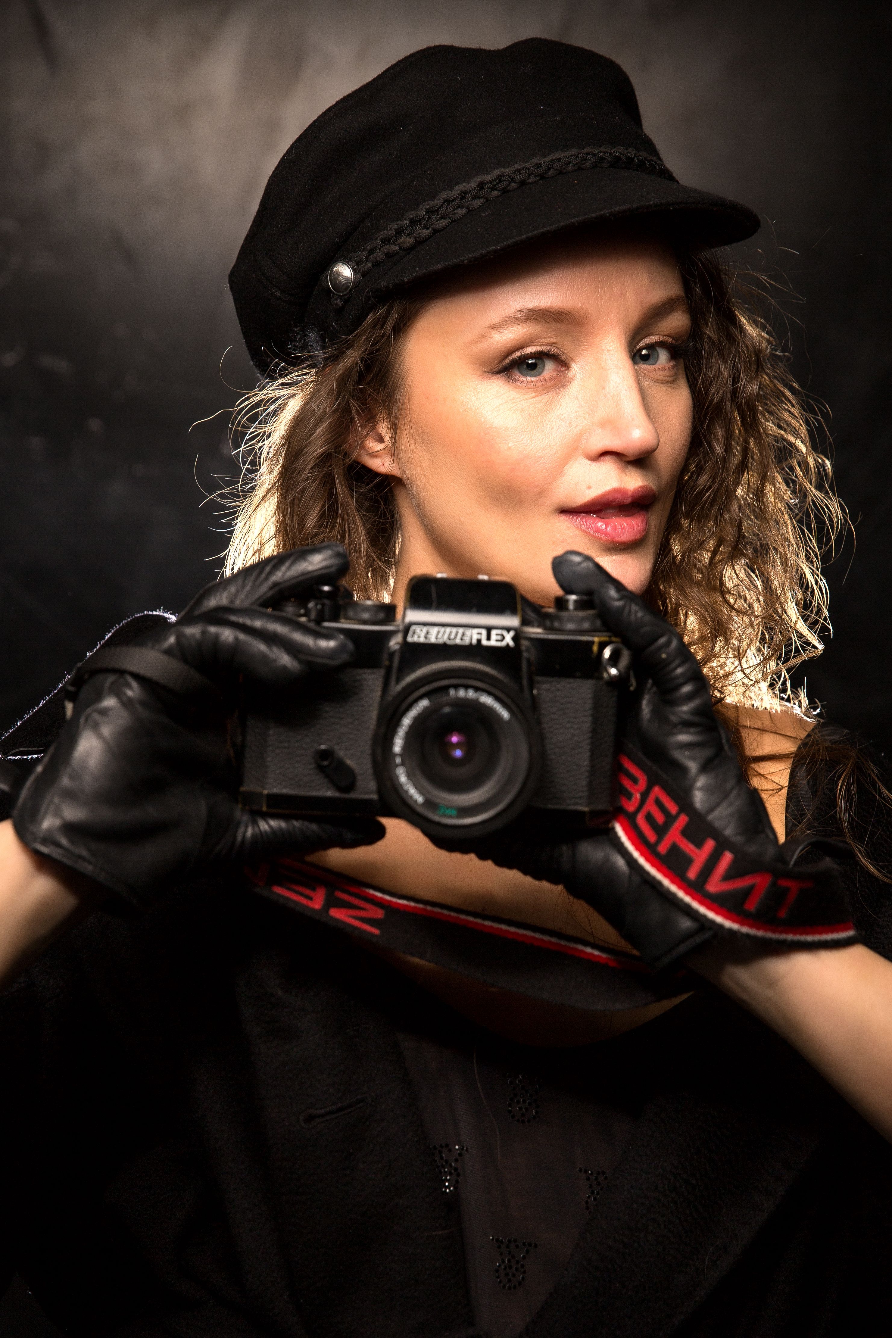 A woman in a black hat and gloves holding a vintage camera, striking a pose.