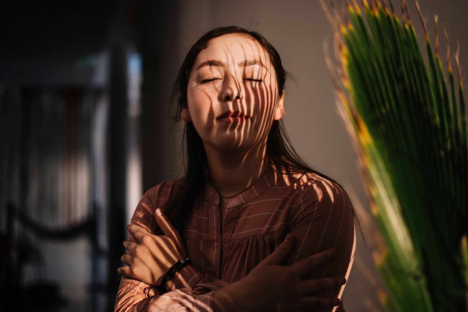 Woman with eyes closed hugging herself as warm sunlight casts striped shadows across her face