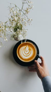 Latte art in a black cup beside delicate white flowers on a pristine table.