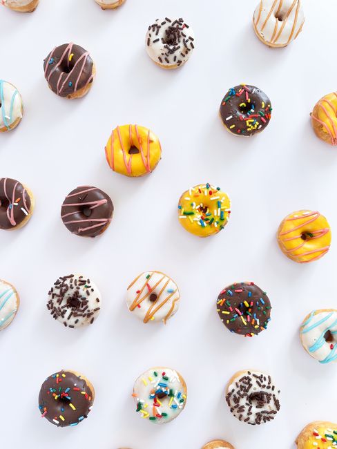 A dozen glazed mini cake donuts covered in rainbow sprinkles, stacked on a wire cooling rack with a bowl of extra sprinkles beside them.