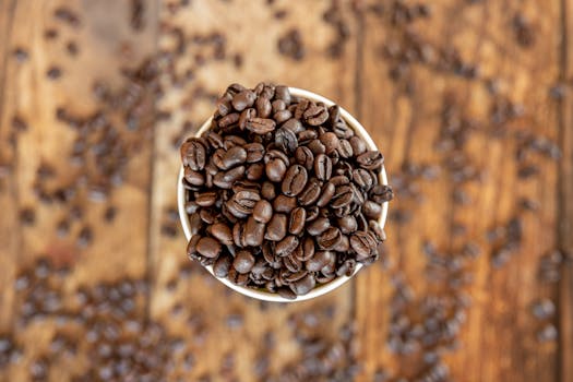 An overhead shot of coffee beans overflowing a cup on a rustic wooden surface.