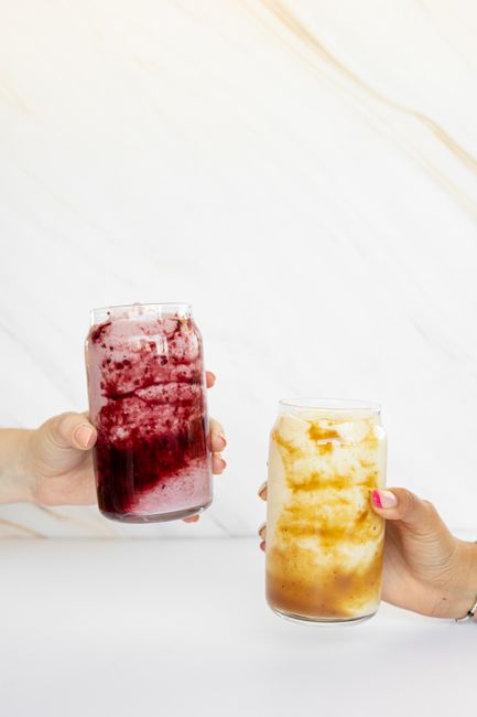 Two hands holding colorful iced drinks in clear glass jars against a bright backdrop.