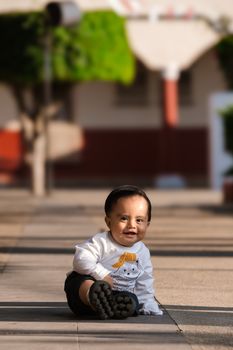 Adorable baby sitting on a sunny outdoor sidewalk, smiling and wearing a cute sweater.