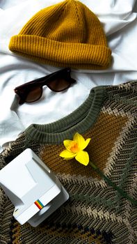 A stylish flat lay featuring a sweater, daffodil, cap, sunglasses, and camera.